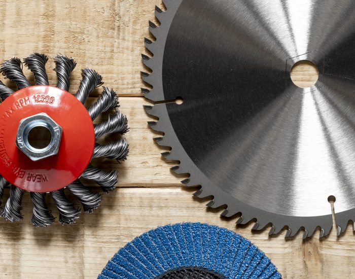 Three circular saw blades lay on a wooden surface. One is a wire brush, another is a toothed saw blade, and the last is a sanding disc.